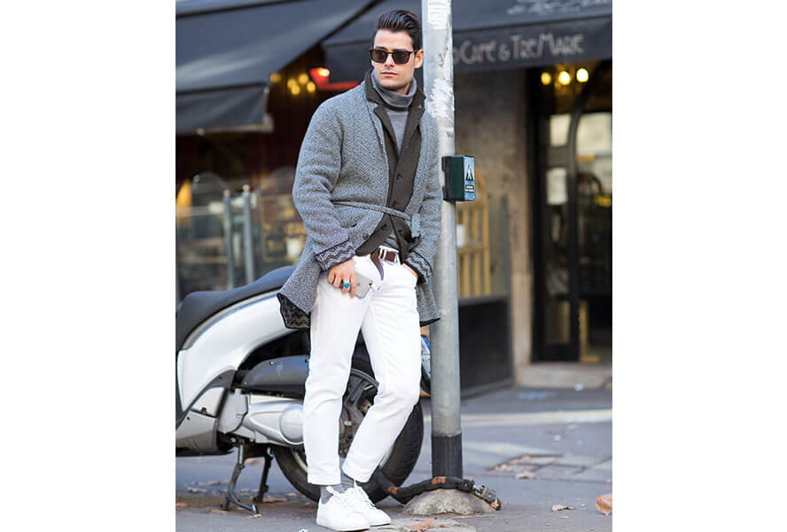 Mann mit weißen Sneakern, einer hellen Hose und grauem Mantel lehnt an einem Strassenschild