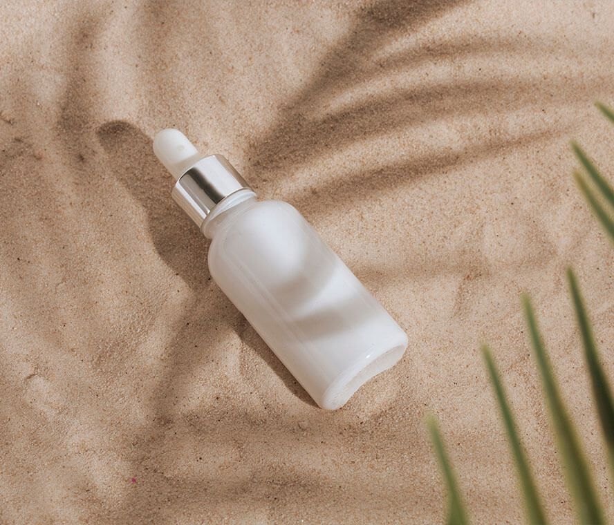Weiße Kosmetikflasche im Sand mit Palmenblatt