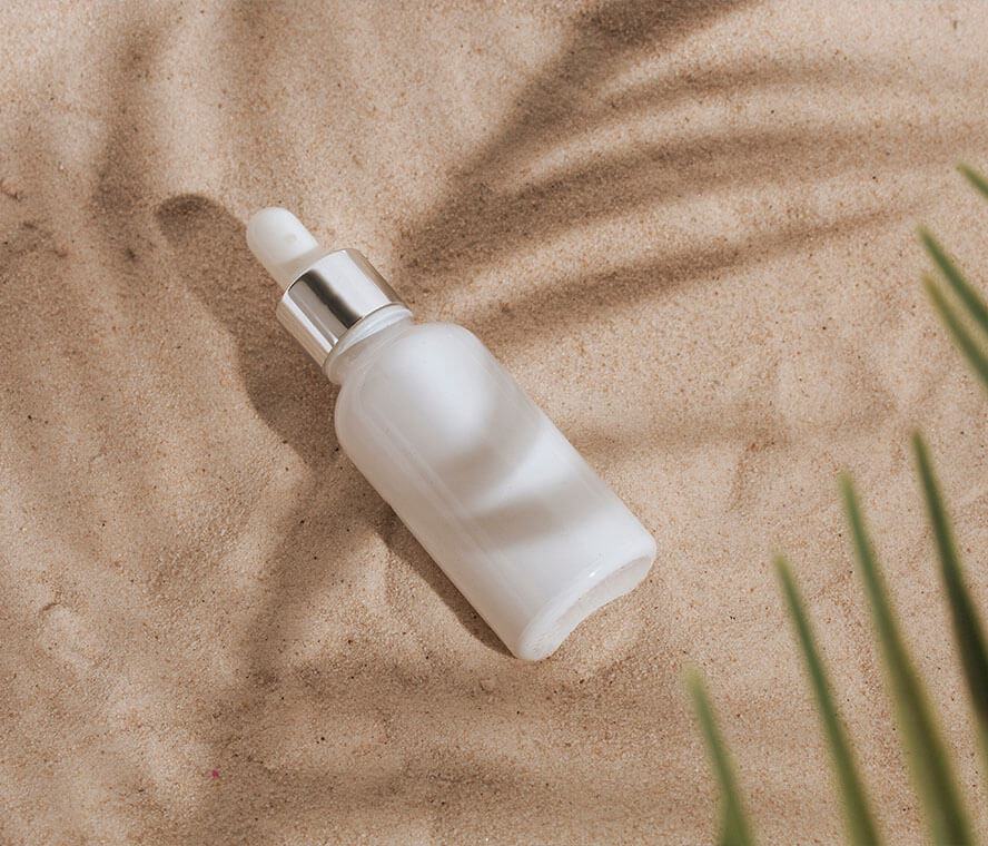 Weiße Kosmetikflasche im Sand mit Palmenblatt