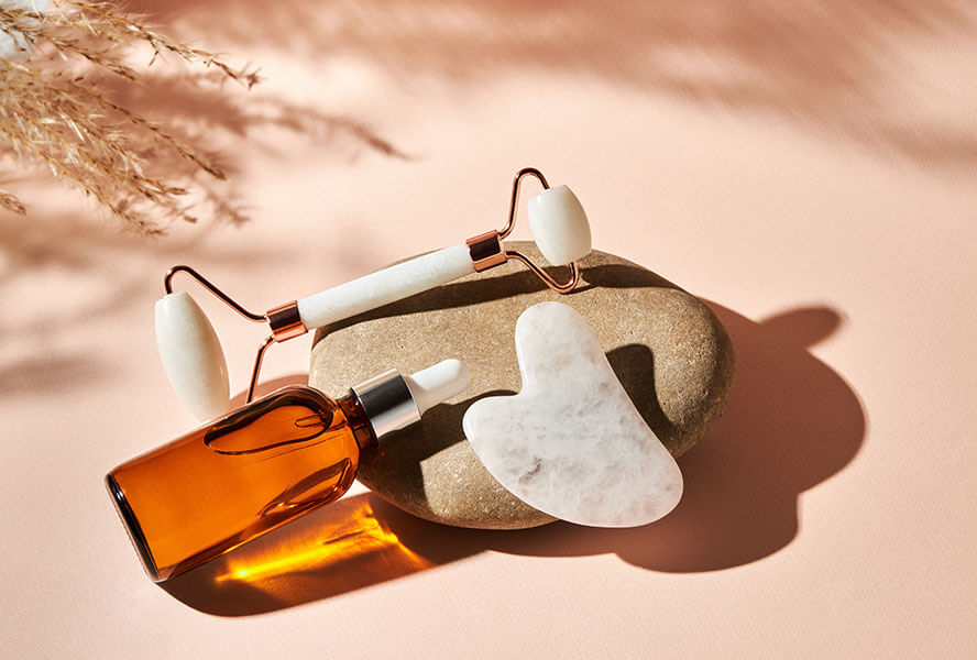 Weisser Gua Sha Stein und Jade-Roller mit pflegendem Gesichtsöl in einer braunen Flasche